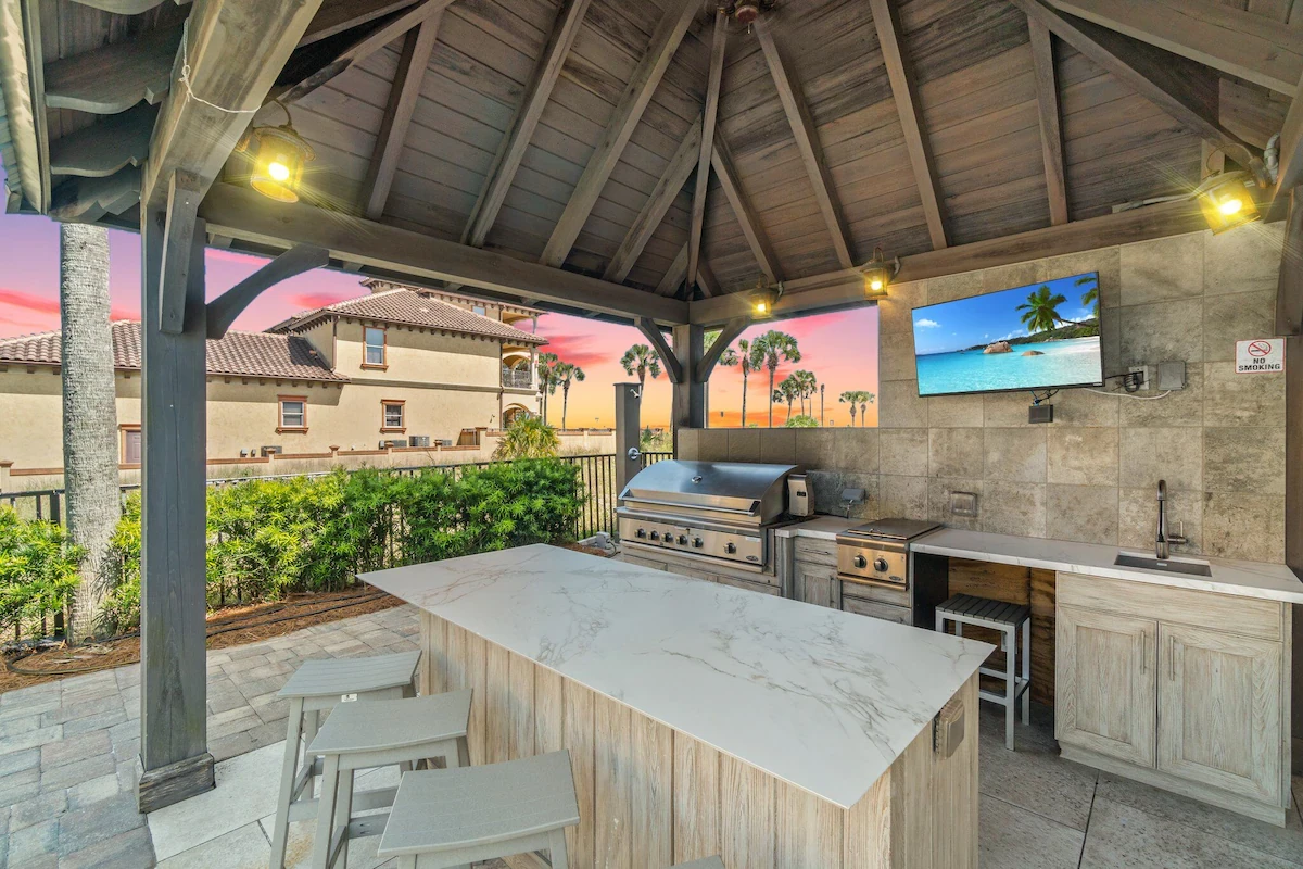 Outdoor kitchen under a wooden gazebo with a grill, bar seating and a TV at sunset